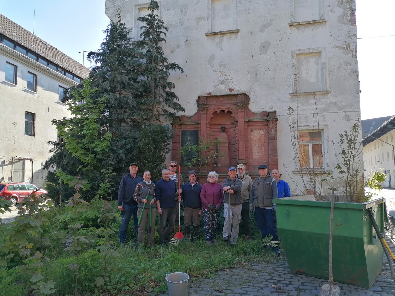 Menschengruppe mit Werkzeug auf der Wiese  vor Haus